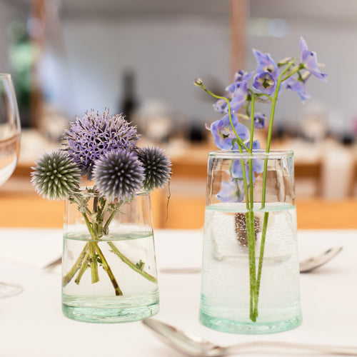 Two clear glass can-shaped vases filled with water and flowers, placed on a white table setting with wine glasses and silverware in a softly blurred dining environment.