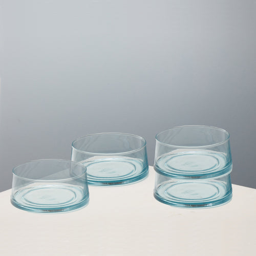 Four clear glass bowls with a light blue tint, arranged on a white surface against a neutral gray background.