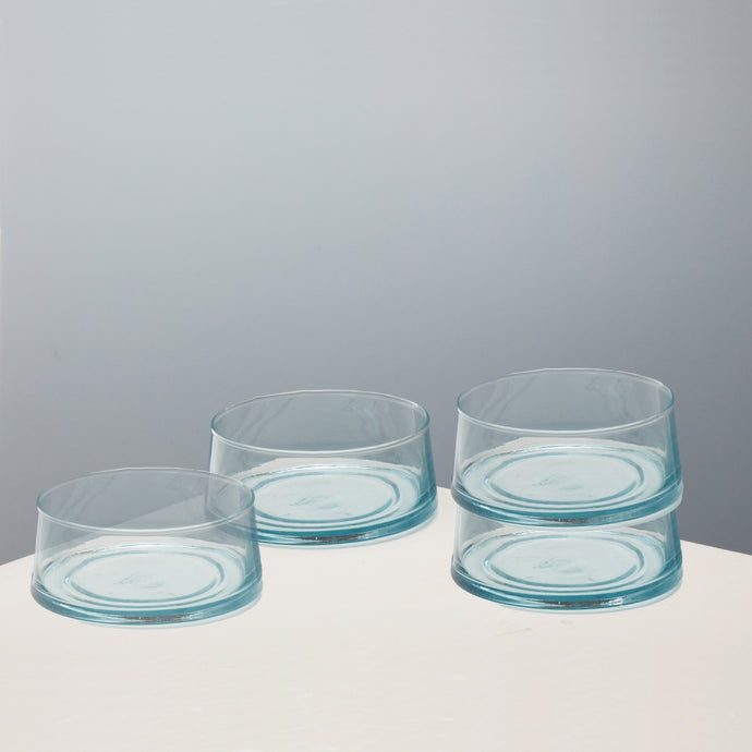 Four clear glass bowls with a light blue tint, arranged on a white surface against a neutral gray background.